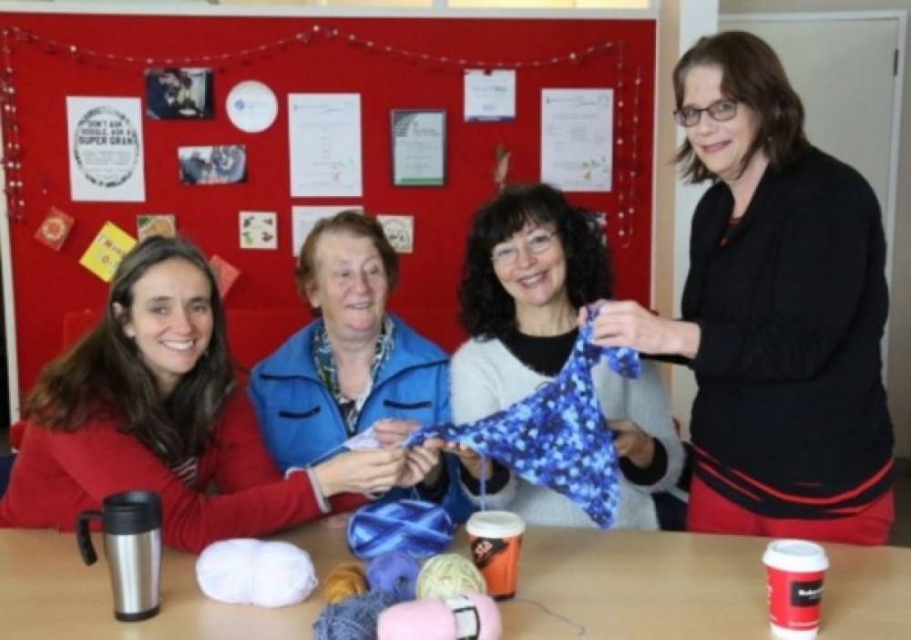 A group of women sit around a large table, knitting or crocheting. There’s an atmosphere of quiet concentration with just a low hum of occasional chat and the odd burst of laughter.